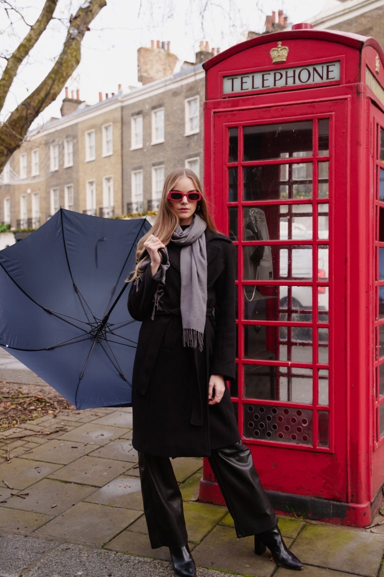 Woman in a dark outfit holding an umbrella beside a red telephone booth