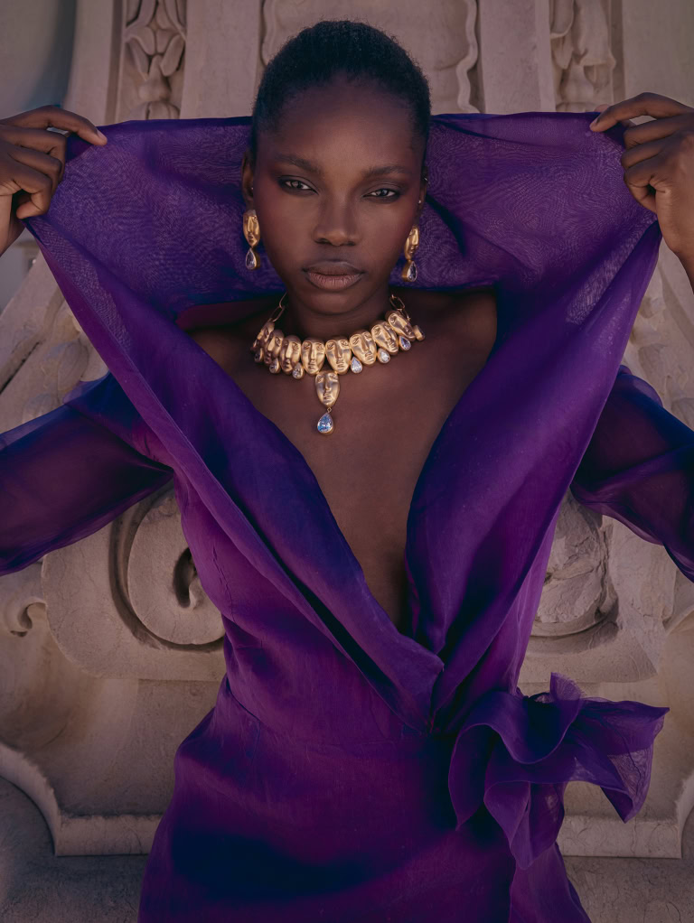Woman holding the collar of a purple blouse open, wearing statement jewelry against a carved stone background.